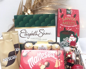 May include: A gift box filled with various treats, including Elizabeth Shaw dark chocolate, Ferrero Rocher, Maltesers, and mini Santa Melts. The box also contains Nescafe coffee packets and a decorative nutcracker. Gold-colored dried plants add a festive touch.