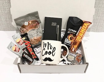 May include: A gift box filled with various items, including a mug with the text "Mr Cool" and a mustache, a black diary with a pen, a chocolate bar, a bottle of body spray, and a bag of cashews.
