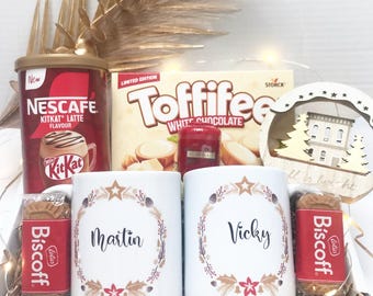 May include: A gift box with a festive theme, containing two white mugs with names, a can of Nescafe KitKat Latte, a box of Toffifee white chocolate, and Biscoff cookies. The mugs have a wreath design with stars and floral accents.