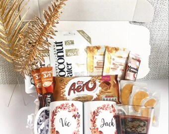 May include: A gift box filled with various treats and items. Includes two white mugs with floral wreaths and names, coffee packets, snacks, eye patches, a candle, and a gold-colored decorative leaf. The box is open, revealing the contents.