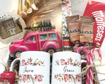 May include: A Christmas-themed gift basket filled with mugs, candy canes, and treats. Two white mugs are decorated with floral designs and the names "Jason" and "Grace". A pink toy car and a small ornament are also included.