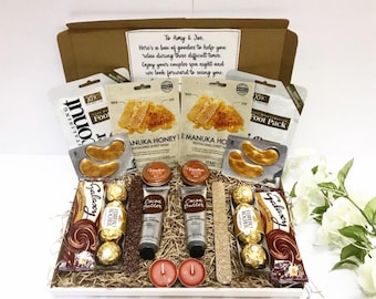 May include: A white gift box filled with spa items, including face masks, eye masks, hand cream, chocolate, and candles. The box is decorated with brown shredded paper and a handwritten note that reads "To Amy & Joe. Here's a box of goodies to help you relax during these difficult times. Enjoy your couples spa night and we look forward to seeing you."