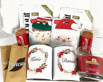 May include: A gift box filled with Christmas-themed items. Includes face masks, coffee packets, personalized mugs with names, Biscoff cookies, and a red candle. The mugs are decorated with a wreath design.
