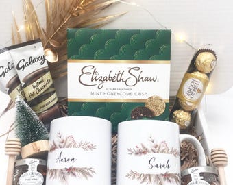 May include: A gift box filled with treats, including two white mugs with names, a box of Elizabeth Shaw dark chocolate, Gala Galaxy instant hot chocolate, Ferrero Rocher chocolates, and jars of honey. Decorative sprigs and a small Christmas tree are also included.