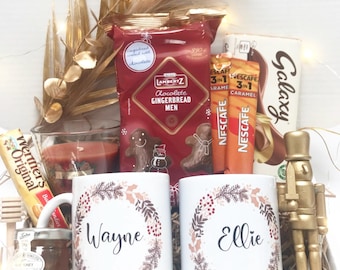 May include: A gift basket filled with treats and mugs. The basket contains two white mugs with names, a package of gingerbread men, coffee packets, a chocolate bar, a jar of honey, and a gold nutcracker.