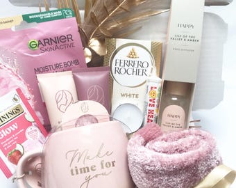 May include: A gift box filled with self-care items. Includes a pink mug with "Make time for you" text, a pink towel, skincare products, a reed diffuser, tea, and a box of chocolates. The box is decorated with gold accents.