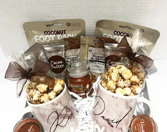 May include: A gift basket filled with self-care items, including two mugs with personalized names, popcorn, a candle, and face masks. The basket is filled with white shredded paper and has a brown ribbon.