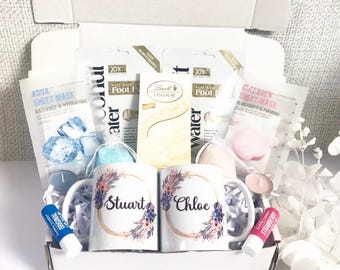 May include: A white gift box filled with self-care items. Includes two floral mugs with names, face masks, foot masks, lip balms, a candle, and a box of chocolates. The mugs have the names "Stuart" and "Chloe" on them.