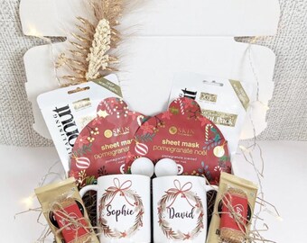 May include: A gift box with Christmas-themed items. Includes two white mugs with names, two sheet masks, two small bags of treats, and decorative elements. The mugs have a wreath design and the names "Sophie" and "David".