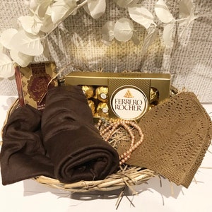 May include: A wicker basket filled with gifts, including a brown scarf, a brown knitted hat, a box of Ferrero Rocher chocolates, a string of pearls, and a small box with Arabic writing.