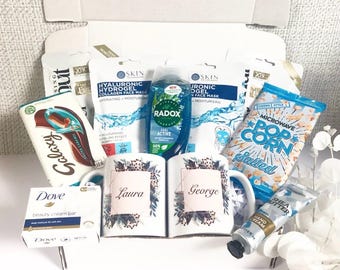 May include: A gift box filled with self-care items. Includes face masks, a bottle of Radox shower gel, popcorn, a chocolate bar, Dove soap, hand cream, and two mugs personalized with names. The mugs have a floral design.