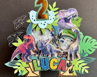 Może przedstawiać: Dekoracja na tort z motywem dinozaurów z zielonym, żółtym, czerwonym i niebieskim banerem "LUCA", numerem 2 i dinozaurami.