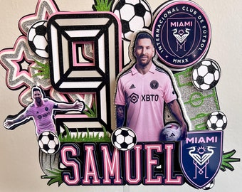May include: A pink and black cake topper with a soccer theme. The topper features a large number 9, a soccer player, soccer balls, and the text "SAMUEL". The topper also includes the logo of Inter Miami CF, a professional soccer club in Miami, Florida.