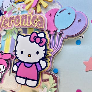May include: A colorful birthday card with a Hello Kitty character, a sun, balloons, and the name Veronica. The card is decorated with glitter and confetti.