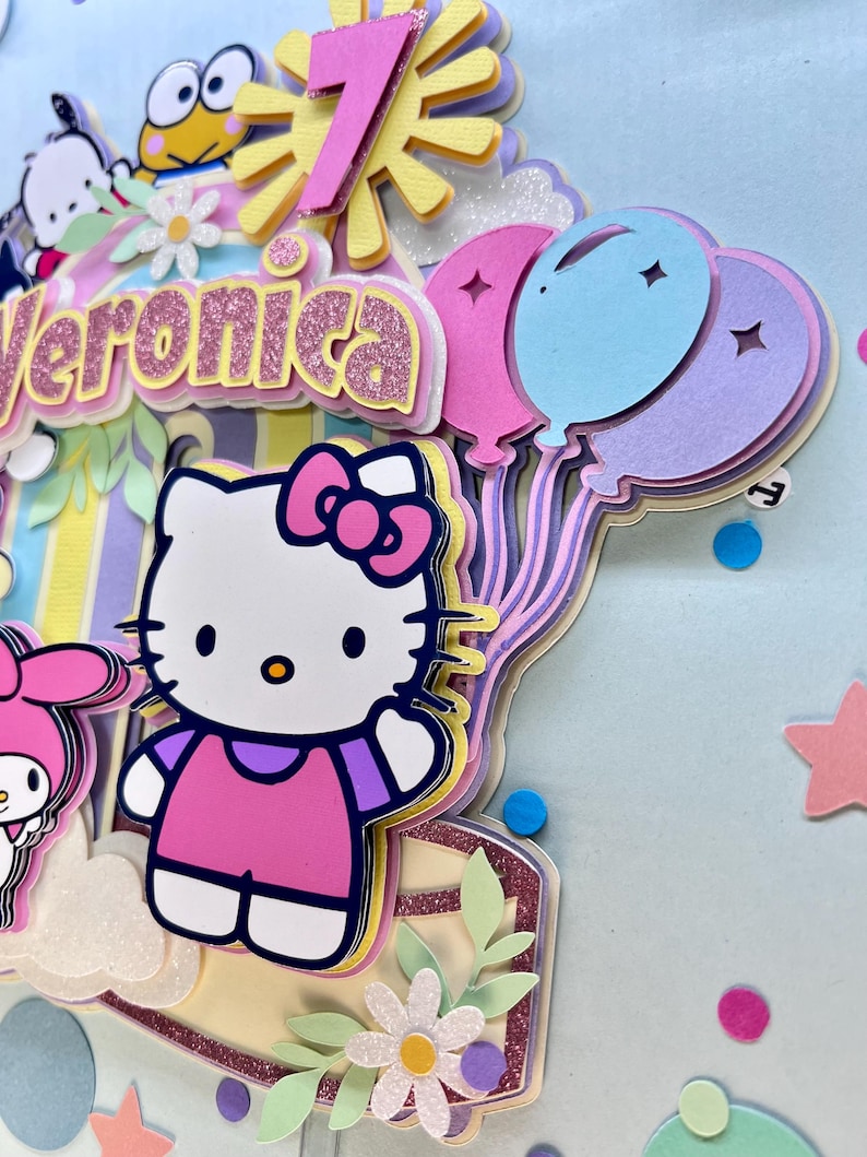 May include: A colorful birthday card with a Hello Kitty character, a sun, balloons, and the name Veronica. The card is decorated with glitter and confetti.