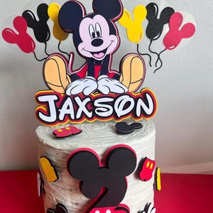 May include: A white frosted cake decorated with a Mickey Mouse theme. The cake topper features a cartoon Mickey Mouse figure, the name "Jaxson", and Mickey Mouse-shaped decorations. The cake is decorated with red, black, yellow, and white accents.