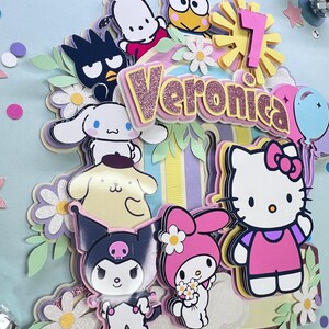 May include: A colorful cake topper featuring Hello Kitty, My Melody, Kuromi, and other popular Japanese cartoon characters. The topper is decorated with flowers and a rainbow-colored background. The name "Veronica" is spelled out in pink glitter letters.