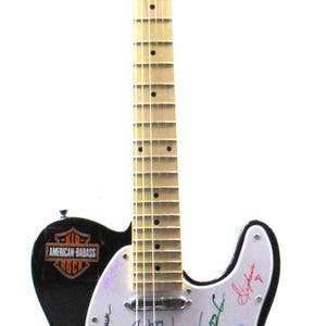 K&ouml;nnte beinhalten: Schwarze E-Gitarre mit wei&szlig;em Schlagbrett, mit einer Grafik eines K&uuml;nstlers und Unterschriften. Die Gitarre hat den Text "AMERICAN-BADASS ROCK" und "LUX PRO" auf der Kopfplatte.
