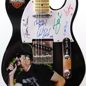 K&ouml;nnte beinhalten: Schwarze E-Gitarre mit einem Musiker-Grafik und Unterschriften auf dem wei&szlig;en Schlagbrett. Die Gitarre hat den Text "Cowboy Baby!" und das Logo "American-Badass Rock". Die Gitarre ist ca. 99 cm lang.