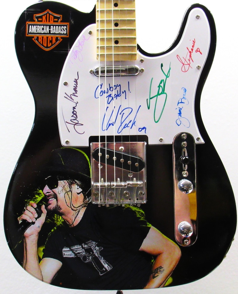 K&ouml;nnte beinhalten: Schwarze E-Gitarre mit einem Musiker-Grafik und Unterschriften auf dem wei&szlig;en Schlagbrett. Die Gitarre hat den Text "Cowboy Baby!" und das Logo "American-Badass Rock". Die Gitarre ist ca. 99 cm lang.