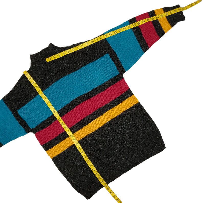 Puede incluir: Un su&eacute;ter negro y gris a rayas con rayas azules, rojas y amarillas. El su&eacute;ter se mide con una cinta m&eacute;trica amarilla.