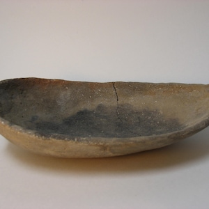 May include: A light brown, oval-shaped ceramic bowl with a crack running down the center. The bowl has a rough, textured surface and a slightly darker brown color on the inside.