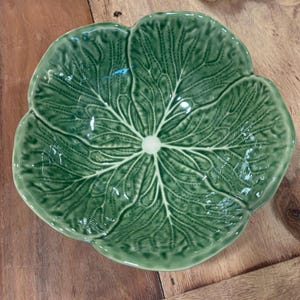 May include: A green ceramic serving dish shaped like a cabbage leaf. The dish has a glossy finish and detailed veining, with a central white accent. The dish is placed on a wooden surface.