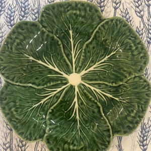 May include: A green ceramic plate shaped like a cabbage leaf. The plate has a glossy finish and detailed veining. The plate is on a patterned tablecloth with blue and white wheat stalks.