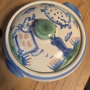 Puede incluir: Una fuente redonda de cerámica con tapa. El plato es blanco con diseños pintados en azul y verde, incluyendo formas florales y abstractas. La tapa tiene un pomo verde y asas azules.