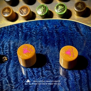 Puede incluir: Dos perillas cilíndricas de madera marrón claro con un gráfico de flor de loto rosa y texto en la parte superior, colocadas sobre el cuerpo de una guitarra de arce flameado azul profundo. Se ven otras perillas borrosas al fondo. Texto: 100% ACTUAL PRODUCT PHOTO.