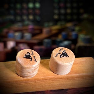 May include: Two cylindrical light-colored wooden knobs with a smooth finish. Each knob features a black graphic logo of a stylized lotus flower and the text "Wants Music" printed on the top surface. The knobs are resting on a rectangular wooden block.