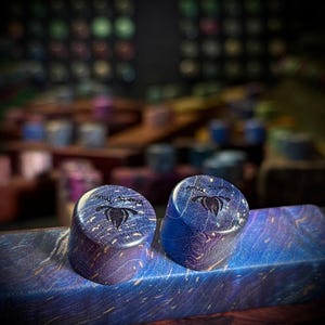May include: Two cylindrical ear plugs with a polished finish, featuring a deep blue and purple wood grain pattern. Each plug is engraved with a black lotus flower icon and the text "Atlantic Music" on the flat top surface.