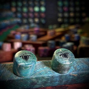 May include: Two cylindrical teal and blue marbled wooden objects with a smooth, polished finish. Each piece features a black logo on the top surface depicting a stylized flower or flame design with the text "Hollow Woods Studio".