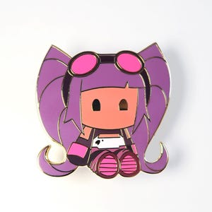 Op de afbeelding: Een kleurrijke emaille pin met een stripfiguur met paars haar, een roze bril en een roze en witte outfit. De pin heeft goudkleurige contouren en is geplaatst tegen een witte achtergrond.