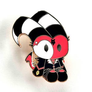 Puede incluir: Un colorido pin de esmalte con un personaje de dibujos animados con una cara roja y blanca, cuernos blancos y negros y un traje negro. El pin tiene detalles dorados y una pequeña cola roja. El personaje está sentado.