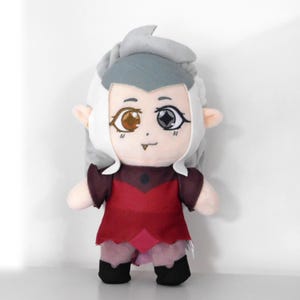 May include: A plush toy of a character with gray hair, a red and purple dress, and one brown and one black eye. The toy has a friendly expression.