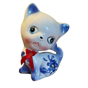 Könnte beinhalten: Vintage Keramik-Katzenfigur mit weißem Körper und blauen Akzenten. Die Katze hat große blaue Augen, rosige Wangen und eine rote Schleife. Blaue Blumenmuster verzieren den Körper und den Schwanz der Katze. Ein bezauberndes Sammlerstück.