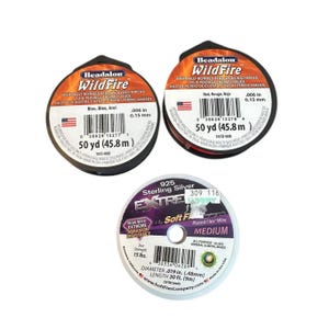 Może przedstawiać: Trzy szpule nici i drutu do nawlekania koralików. Dwie szpule WildFire termicznie wiązanej nici do tkania koralików w kolorze niebieskim i czerwonym, każda po 45,8 m. Jedna szpula posrebrzanego drutu flex 925, średnica 0,48 mm, długość 9 m.