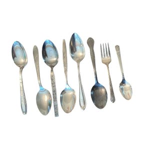 Puede incluir: Un surtido de cubiertos antiguos, que incluye cucharas y un tenedor, dispuestos sobre un fondo blanco. Las cucharas varían en tamaño y diseño, algunas con patrones ornamentados. El tenedor tiene cuatro púas.