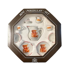 May include: A miniature porcelain tea set in a brown octagonal box. The set includes a teapot, creamer, sugar bowl, cups, and saucers. Each piece is white with a red trim and a teddy bear design. The box reads "MINI - PORZELLAN - SET".