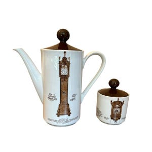 May include: White ceramic teapot and sugar bowl set with brown accents. The teapot features a long spout and handle, with a brown lid. Both pieces are decorated with a brown illustration of a grandfather clock and text.