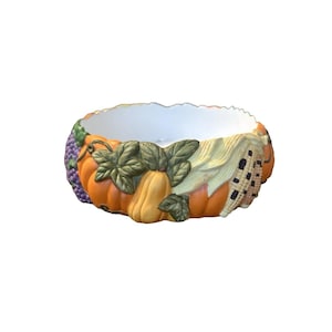 Op de afbeelding: Een keramische kom met een wit interieur en een buitenkant versierd met herfstelementen. De kom heeft oranje pompoenen, gele kalebassen, groene bladeren, paarse druiven en maïs. De kom is een decoratief stuk.