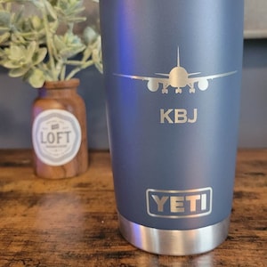 May include: A blue Yeti Rambler tumbler with a stainless steel lid and bottom. The tumbler has a laser-etched design of an airplane with the initials "KBJ" underneath. The Yeti logo is also laser-etched on the bottom of the tumbler.