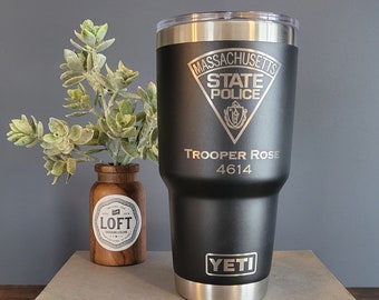 Könnte beinhalten: Ein schwarzer Yeti-Becher mit einem silbernen Deckel und einem lasergravierten Design. Das Design zeigt das Logo der Massachusetts State Police, die Worte "Trooper Rose" und die Nummer "4614".