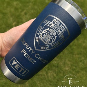 May include: A navy blue stainless steel tumbler with a silver rim and base. The surface features an engraved New York City Police Department badge, the text "DEPUTY CHIEF PEREZ," and the YETI brand logo. The design is etched into the powder-coated finish.