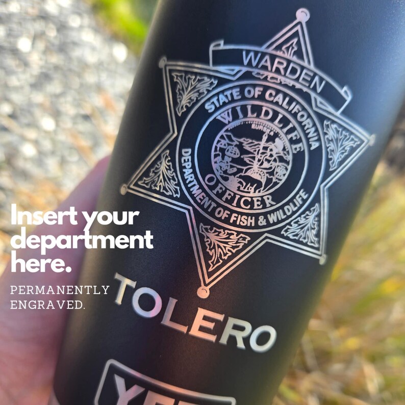 May include: Black stainless steel tumbler with a silver engraved design of a California State Wildlife Officer badge. The text "WARDEN STATE OF CALIFORNIA WILDLIFE DEPARTMENT OF FISH & WILDLIFE OFFICER" is engraved around the badge. The text "TOLERo" is engraved below the badge.