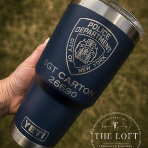 May include: A navy blue YETI tumbler featuring a laser-engraved New York City Police Department shield, the text SGT CARTON, and the number 26690. The stainless steel cup has a clear lid and a matte finish, with the YETI logo at the base.