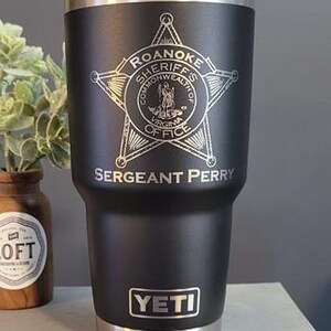 May include: A black Yeti tumbler with a silver lid and base. It has a laser engraved design of a star with the words "Roanoke Sheriffs Commonwealth of Virginia Office" and "Sergeant Perry".