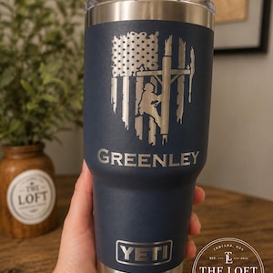 May include: A navy blue stainless steel tumbler with a clear lid, featuring a laser-engraved design of an American flag combined with a utility pole worker silhouette. The name GREENLEY is engraved below the graphic, with the YETI logo at the base.