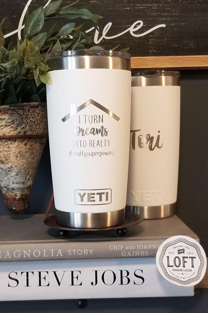 May include: Two white Yeti tumblers with silver accents. One tumbler has the text "I TURN Dreams INTO REALTY #realtysuperpowers" and the Yeti logo. The other tumbler is inscribed with the name "Tori".